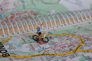 Auf einer aufgeschlagenen Landkarte mit Bonn umrandet steht ein Miniaturradfahrer und reisst die Arme hoch.