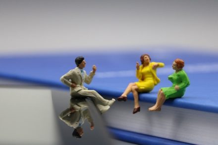 Drei Miniaturfiguren, eine männlich und zwei weiblich, sitzen auf einem Buch und einem E-Reader gegenüber und scheinen zu plaudern